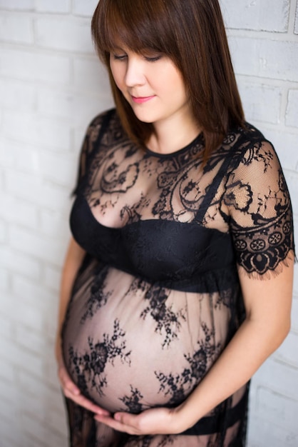 Close up portrait of pregnant woman in black dress touching her belly over white brick wall