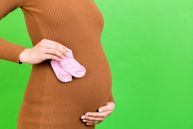 緑の表面で女の赤ちゃんのためのピンクの靴下を保持している茶色のドレスを着た妊婦の肖像画をクローズアップ