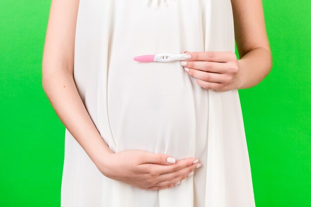 Close up portrait of pregnant woman in white dress holding positive pregnancy test against her belly at green background. Healthy pregnancy. Copy space.