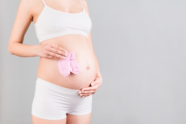 Close up portrait of pregnant woman in white underwear holding pink socks for a baby girl at gray background. Waiting for a child. Copy space.