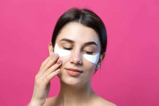 Close up portrait of pretty attractive girl with naked shoulders using patches under eyes. Standing over pink background.