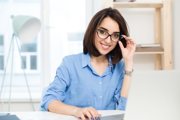 Close-up portrait of a pretty brunette girl sitting at the table in office. She wears blue shirt and holds glasses. She is smiling to the camera.