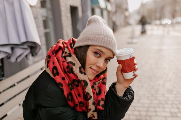 Close up portrait of romantic pretty lady with great make up wearing beige knitted cap and red scarf is drinking coffee outdoor on summer terrace in the city