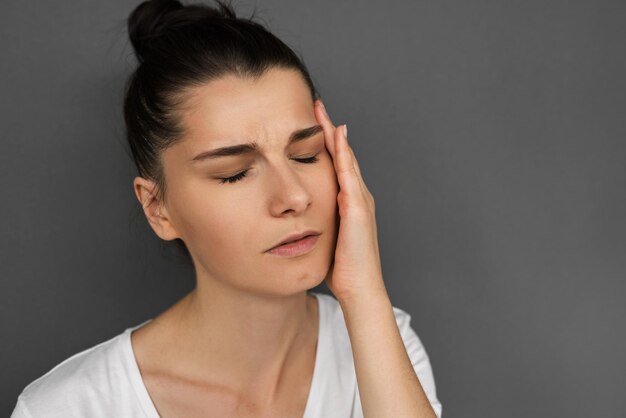 Close up portrait of sad young brunette female suffering from bad headache or migraine while facing stress at work with closed eyes have painful expression touching temple with hand on gray