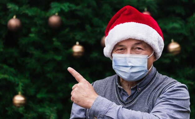 Close up ritratto di uomo anziano che indossa un cappello di babbo natale e mascherina medica con emozione e mostra il dito. sullo sfondo di un albero di natale. pandemia di coronavirus