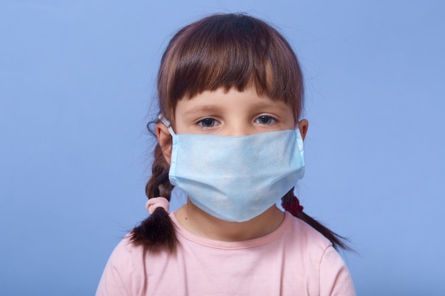 Close up portrait of serious sweet little girl wearing medical mask, looking directly ar camera