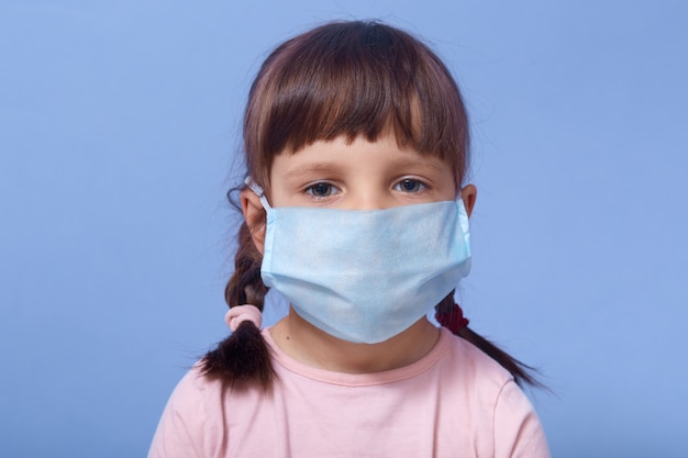 close up portrait of serious sweet little girl wearing medical mask, looking directly ar camera