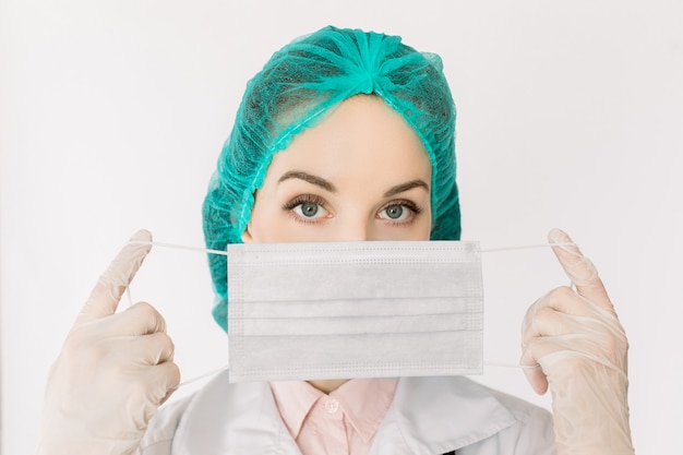 Close-up portrait of serious young nurse or doctor in cap, gloves and white coat, showing how to wear medical face mask, isolated on white background. Deseases and infections prevention