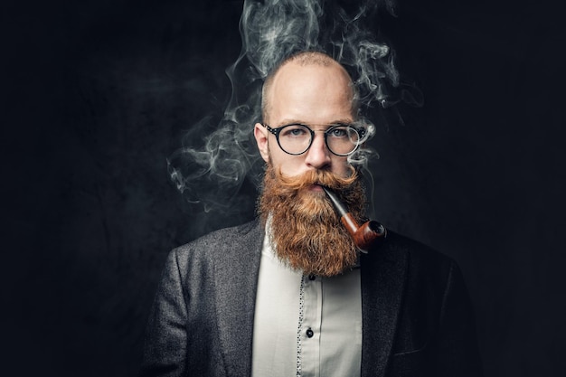 Close up portrait of shaved head aristocratic male in eyeglasses smoking pipe over grey background.