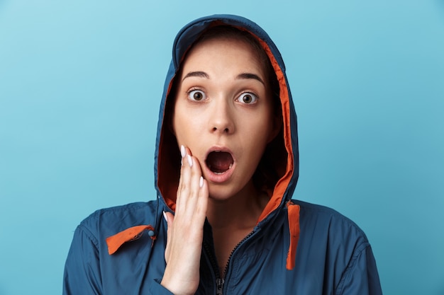 Close up portrait of a shocked young girl wearing hood standing isolated over blue wall