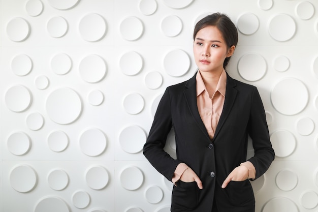 Крупным планом портрет азиатской бизнес-леди, стоящей, улыбающейся и кладущей руку в сумку костюма, демонстрирующую удобство фотографирования на фоне с множеством белых круглых поверхностей.