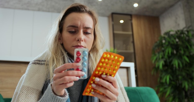 Close up of portrait of sick woman in warm plaid sitting on sofa at home and choosing medicine. Infected female holding pills in hands indoors. Concept of healthcare, illness and flu.