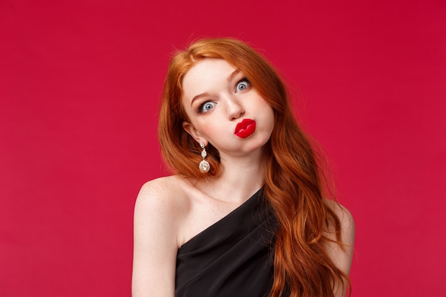 Photo close-up portrait of silly and funny pretty redhead woman in red lipstick, makeup and black evening dress, tilt head pouting like wish and fold lips mwah, stand on a red wall