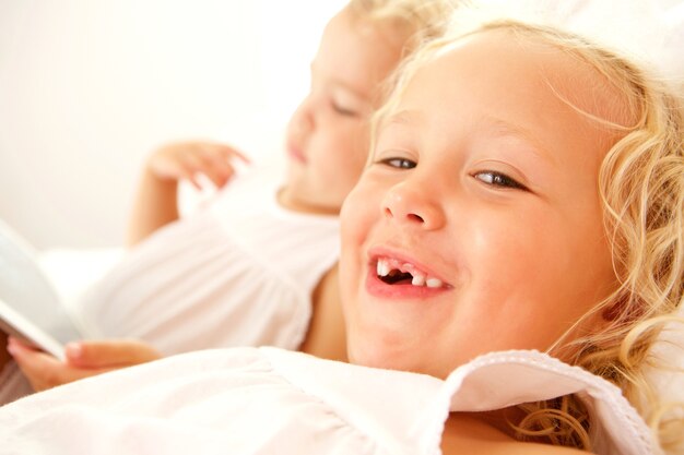 Close up portrait of smiling adorable girl on bed with sister using digital tablet 