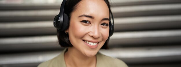 Photo close up portrait of smiling asian girl in headphones listens to music outdoors looking happy