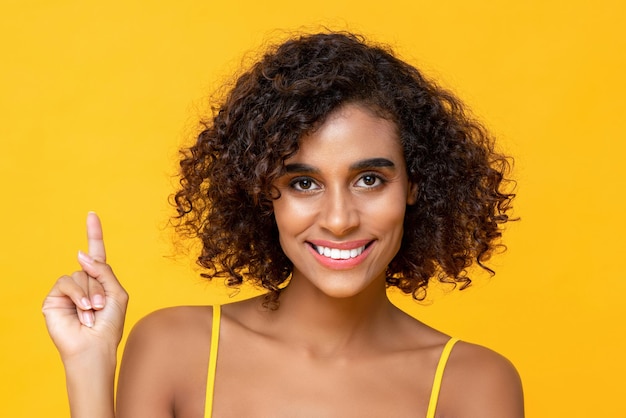 노란색 배경에 격리된 한 손가락을 가리키며 카메라를 바라보며 웃고 있는 아름다운 아프리카계 미국인 여성의 초상화를 클로즈업