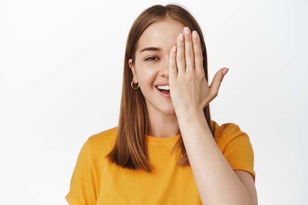 Close up portrait of smiling blond girl, cover half of face and laughing, looking at camera with one eye, checking vision in eyewear store, standing against white background