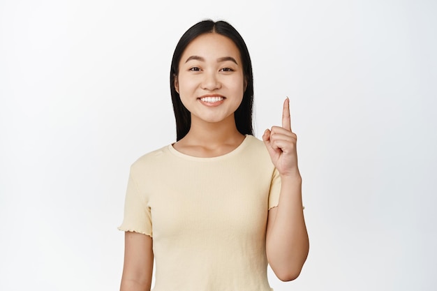 Close up portrait of smiling cute asian girl pointing fingers up showing advertisement standing in yellow tshirt over white background