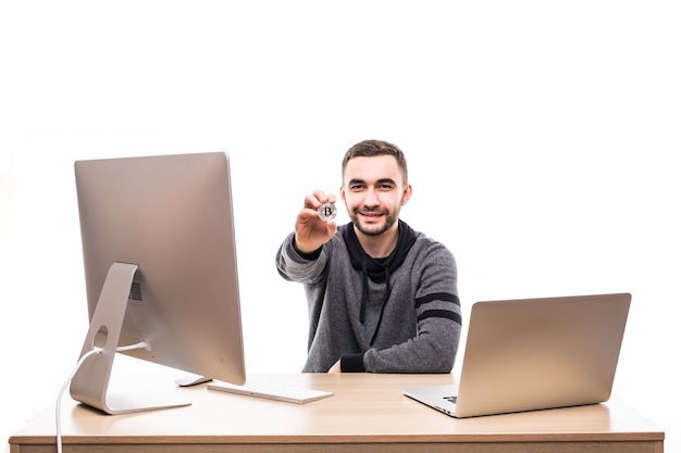 Close up portrait of a smiling entrepreneur holding bitcoin while sitting at desk with laptop and computer isolated over white