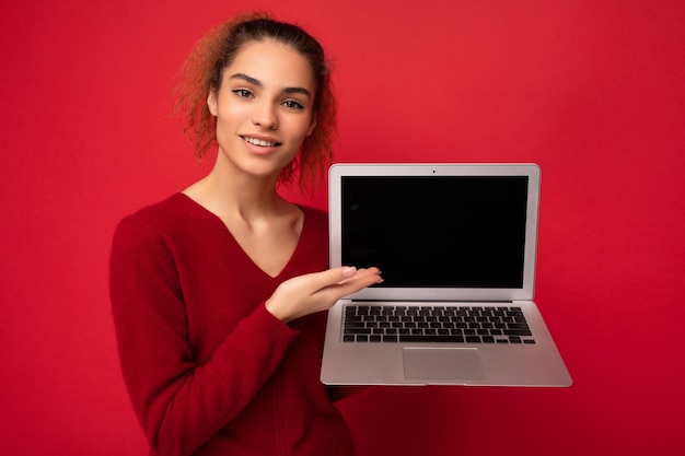 Ritratto del primo piano di sorridere felice bella donna bionda scura che tiene il computer portatile che guarda l'obbiettivo che mostra alla tastiera del netbook che indossa un maglione rosso isolato sopra priorità bassa rossa della parete.