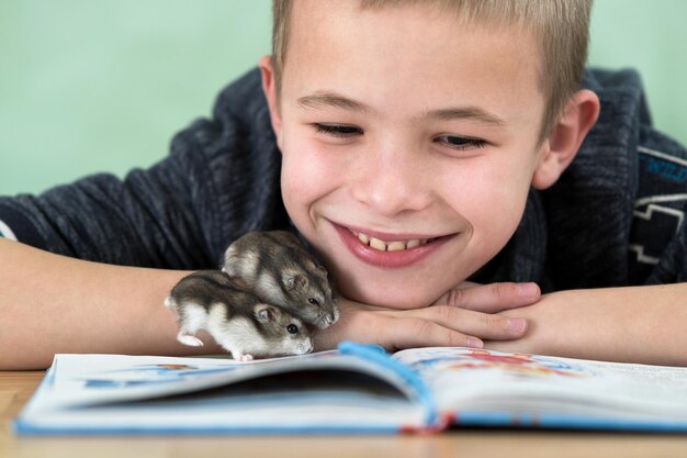 Close up portrait of smiling little boy reading book with small pet hamsters. Concept of studying with good mood.