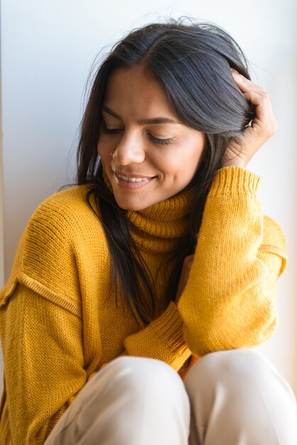 Close up portrait of a smiling lovely young woman dressed in sweater sitting at the window indoors, eyes closed