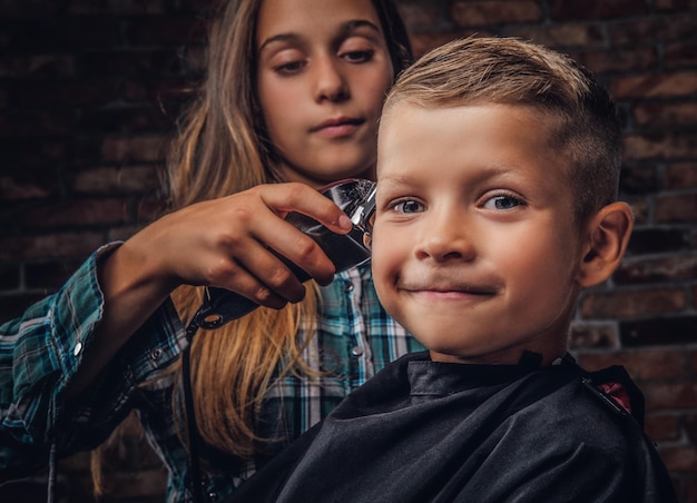 Close-up portrait of a smiling preschooler boy getting haircut. The older sister cuts her little brother with a trimmer against a brick wall.