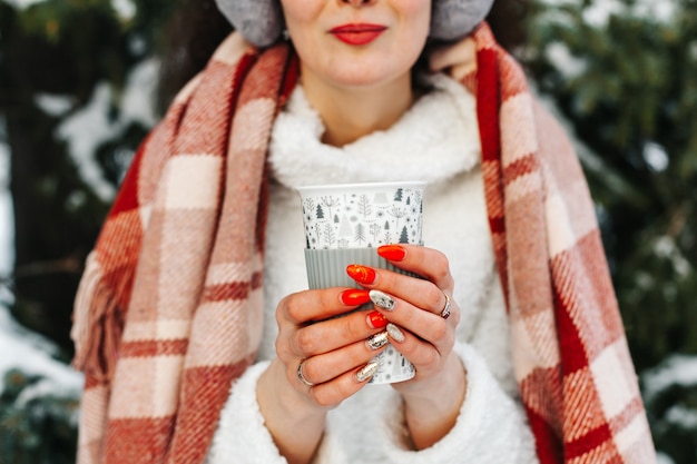 겨울 숲에서 뜨거운 코코아 한 잔을 들고 웃는 젊은 여성의 초상화를 닫습니다