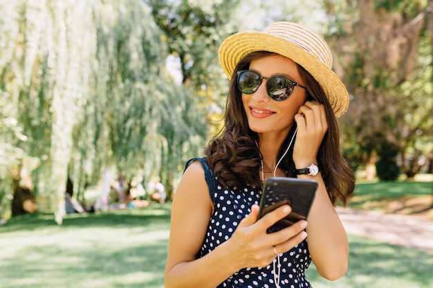 Close up portrait of style woman is walking in the summer park wearing summer hat and black sunglasses and cute dress. She is listening music.