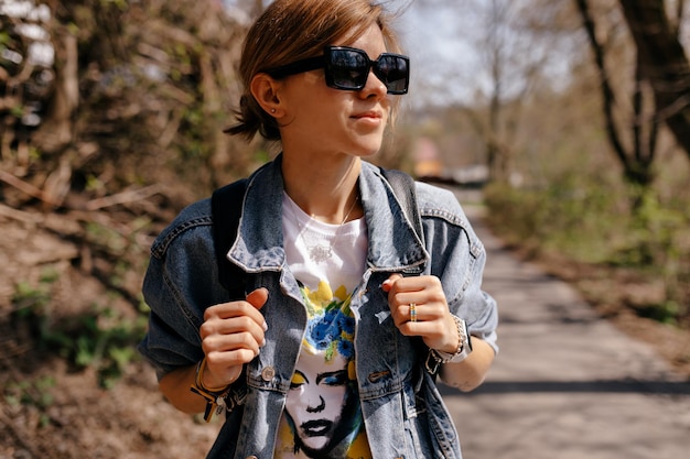 Крупный план портрета стильной девушки со светлыми волосами в солнцезащитных очках и джинсовой куртке, с улыбкой отводящей взгляд в солнечном свете на фоне леса