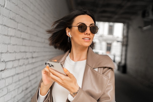 Foto close up ritratto di elegante donna dai capelli corti con gli auricolari in cappotto di pelle casual e occhiali da sole utilizzando smartphone e posa sopra il muro di mattoni urbano