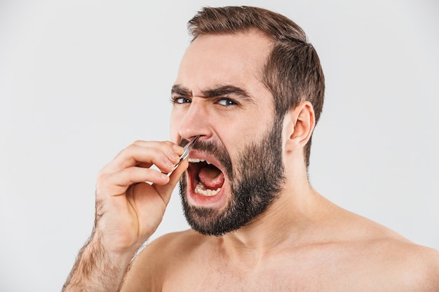 Close up portrait of a suffering bearded man standing isolated over white, plucking out his nose hair