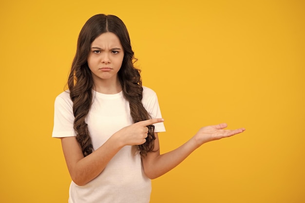 Close up portrait of teenager child girl showing at copy space\
pointing to ads advertising isolated over yellow background unhappy\
sad teenager girl