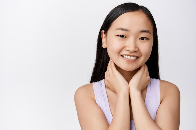 Close up portrait of tender asian girl with soft natural skin without makeup touching neck and smiling cute standing over white background