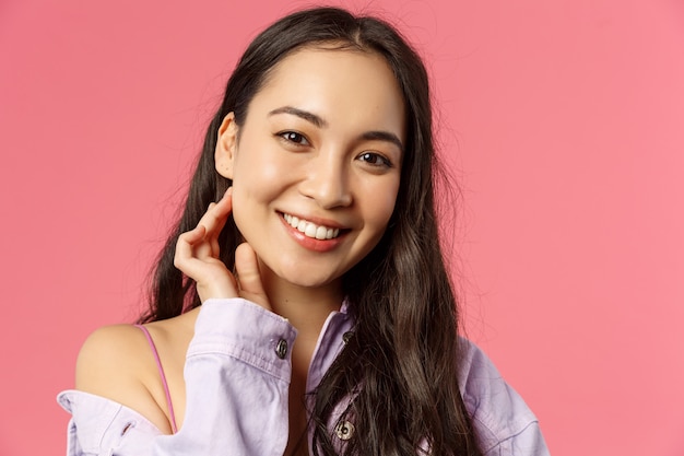 Photo close-up portrait of tender, beautiful young asian woman with clean skin, no pimples or blemishes, smiling lovely camera, touching neck and look camera romantic, stand pink wall