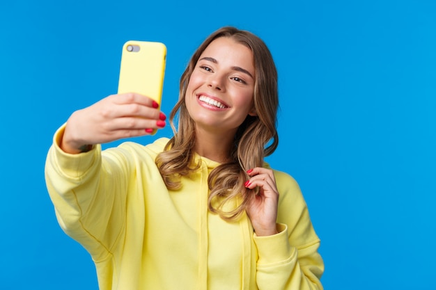 Close-up portrait tender cute blond girl in yellow hoodie, holding mobile phone, taking selfie with smartphone posing adding photo filter in appllication, standing 