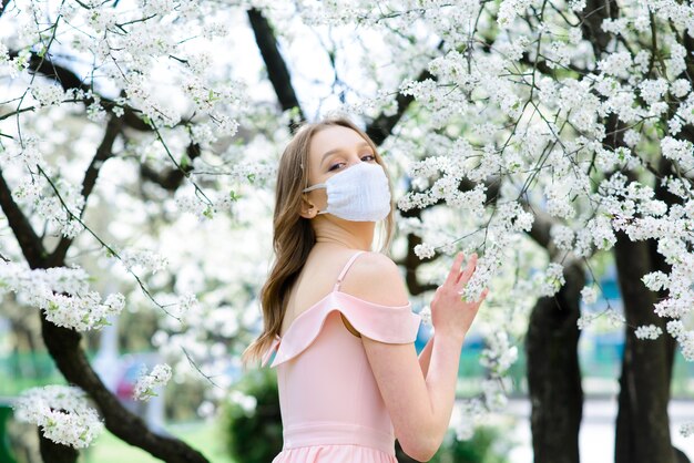 Close up portrait of tender girl under a blossoming cherry tree with a mask from the coronavirus.