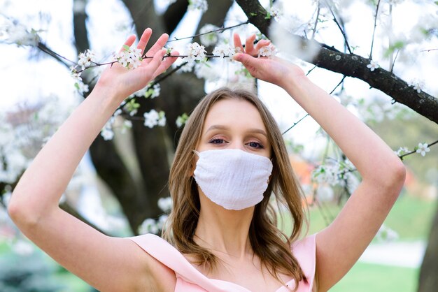 Close up portrait of tender girl under a blossoming cherry tree with a mask from the coronavirus.