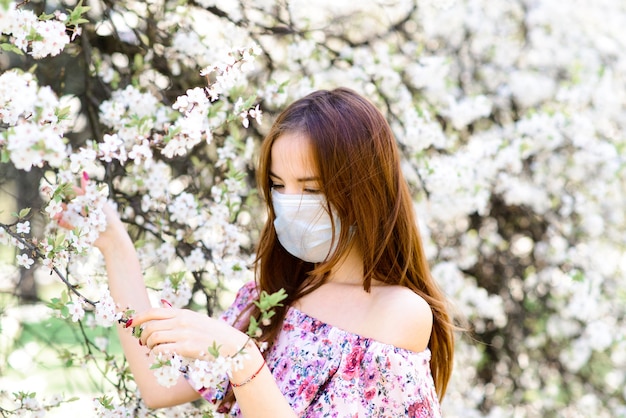 Close up portrait of tender girl in a white blouse under a blossoming cherry tree with a mask with flowers on from the coronavirus.