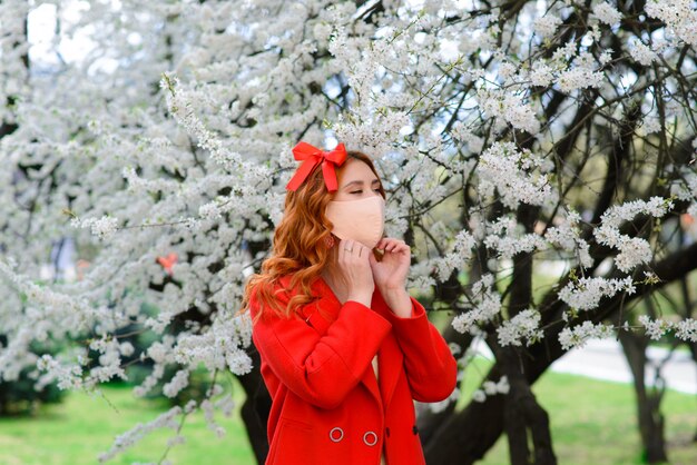 Close up portrait of tender red-haired girl in a red coat under a blossoming cherry tree with a mask with flowers on from the coronavirus.