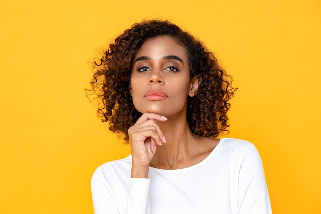 Close up portrait of thinking young African American woman looking at camera with one hand touching chin on studio yellow studio background