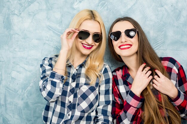 Close-up portrait of two female friends in checkered shirts, jeans and sunglasses on the blue painted wall background