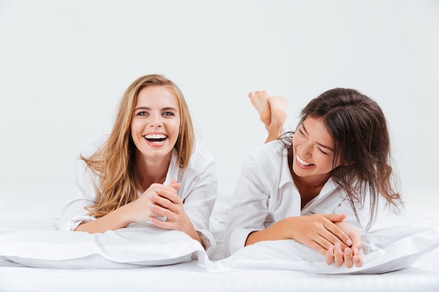 Close up portrait of two laughing beautiful woman lying together on bed with white sheets