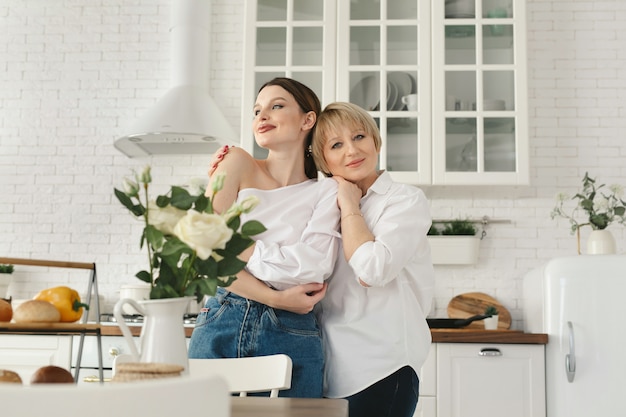 Close-up portrait of two nice attractive lovely tender cheerful women mom mommy adult daughter spending time day together in white light interior house flat apartment indoors