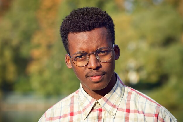 Close up portrait of unhappy serious angry disappointed annoyed dissatisfied black guy, young handsome African Afro American ethnic man in shirt and glasses looking at camera outdoors in autumn park