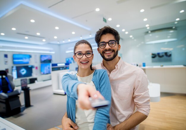 Close up portrait view of cute excited charming young student\
girl with eyeglasses holding tv remote control while her boyfriend\
hugging her in a tech store.