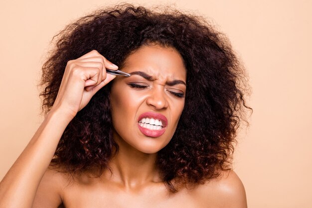 Close-up portrait of well-groomed irritated girl using metal forceps eyebrow