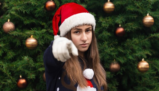 Close up Portrait of woman wearing a santa claus hat with emotion and shows a finger at the camera. Against the background of a Christmas tree