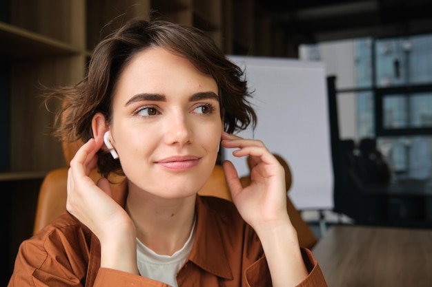 Close up portrait of woman in wireless earphones smiling and sitting in office listening to music
