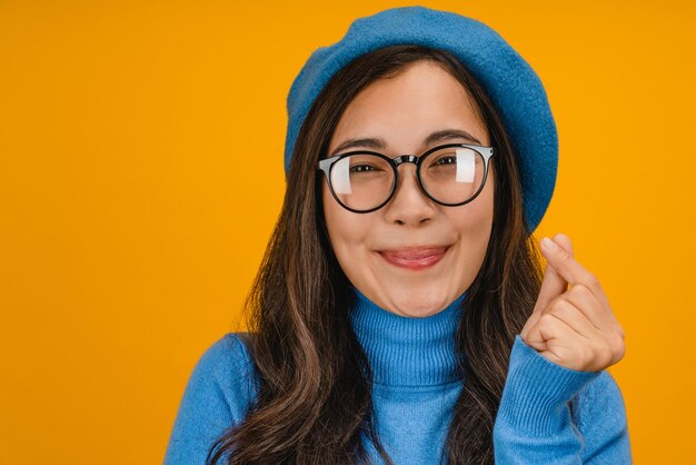 Close up portrait of woman with dark hair makes korean love sign isolated over yellow background